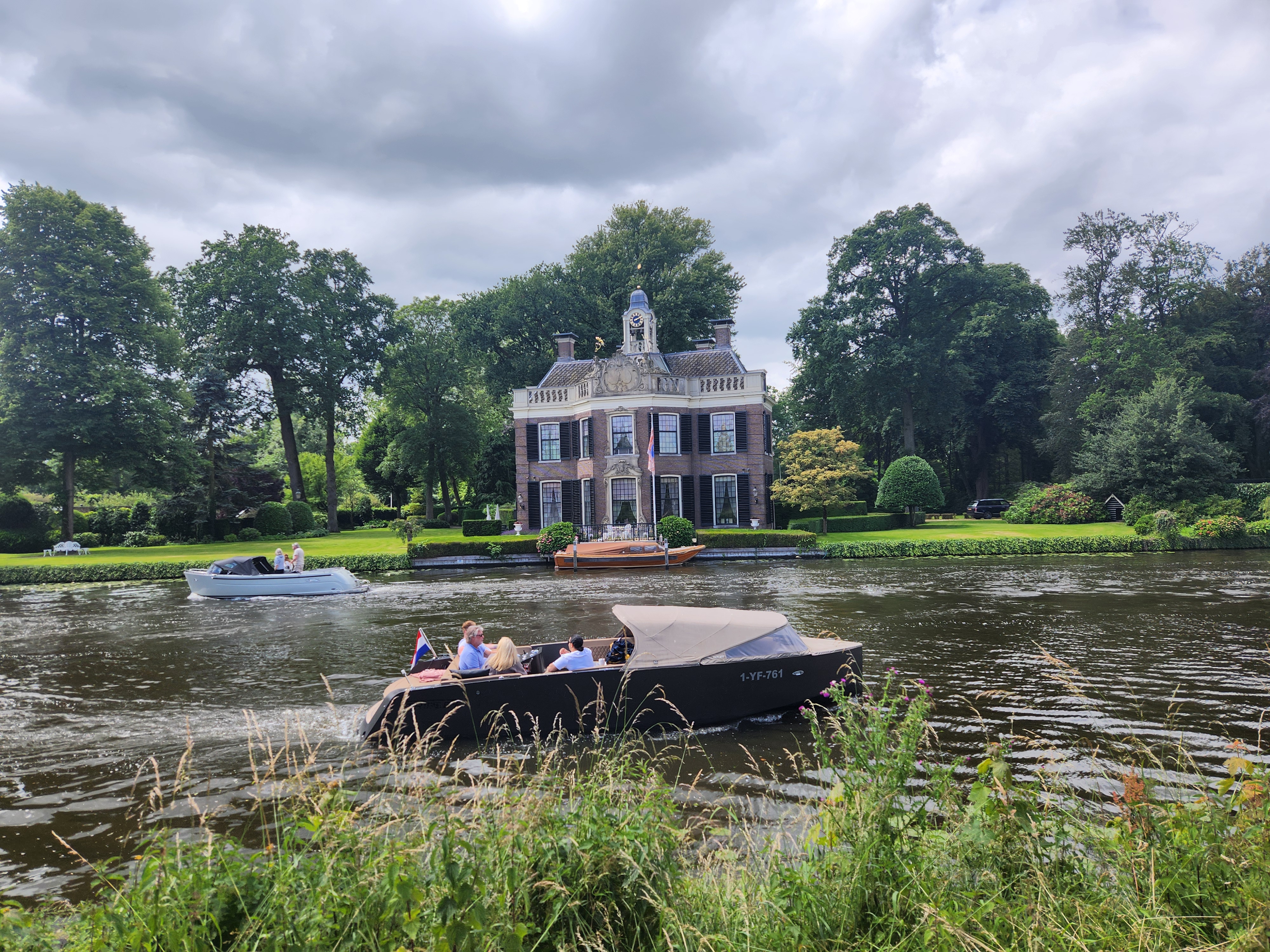 A very posh looking river home in front of the river. In the river is a very posh looking river cruiser with very posh looking people looking like they are having a good time